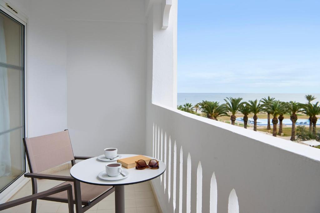 Balkons ar skatu uz jūru, uz galda kafija, grāmata un saulesbrilles. Apkārt palmām un baseinam.