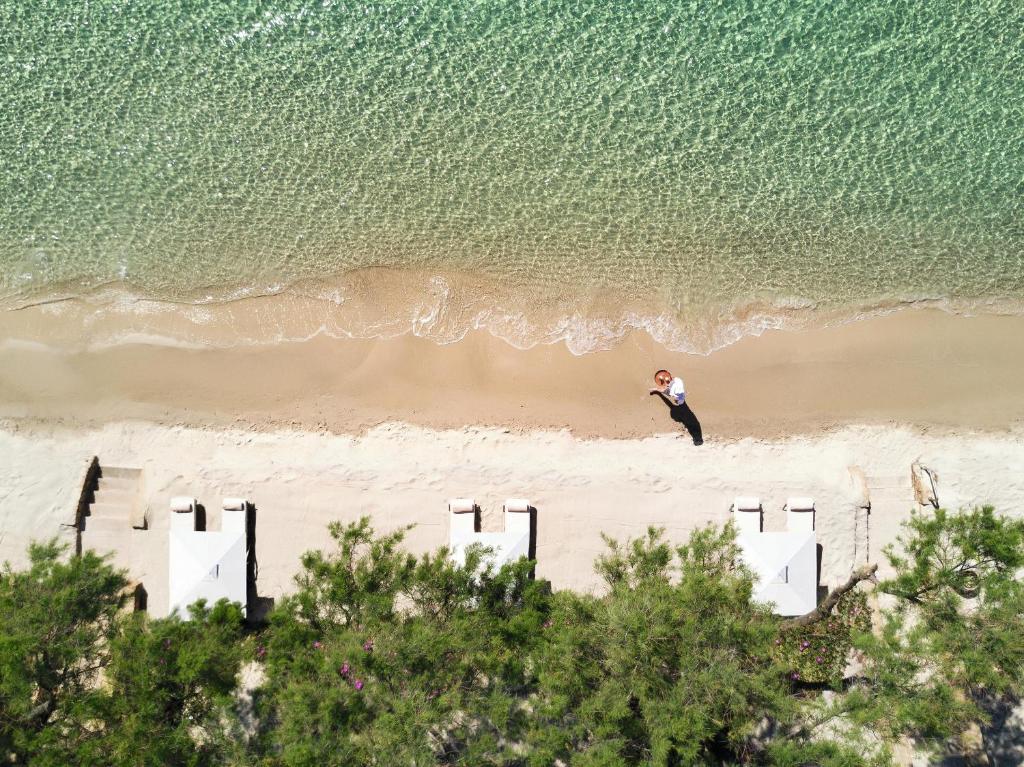 Pludmales skats no augšas, kur redzama cilvēka siluets, kas staigā pa smilšu krastu blakus dzidram ūdenim. Apkārt ir saulessargi un zaļumi, radot mierīgu atpūtas atmosfēru.