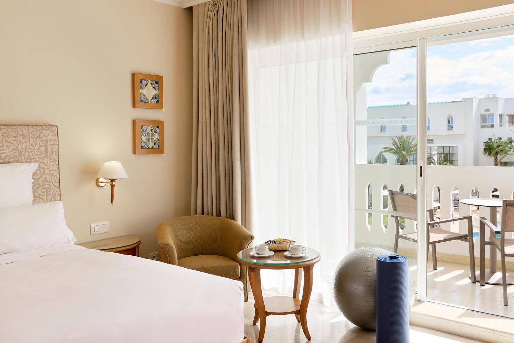 Mūsdienīga viesnīcas istaba ar ērtu gultu, dīvānu un galdu, kurā ir kafijas tase un uzkodas, lieliem logiem un balkonu, kas sniedzas uz brīnišķīgu skatu.