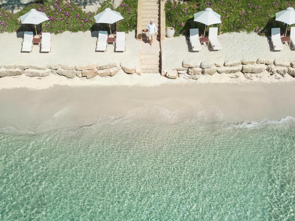 Pludmales ainava ar saulessargiem un sauļošanās krēsliem, kas izvietoti gar krastu, un tīru, dzidru ūdeni. Aiz krasta redzama zāle ar ziediem un akmeņiem.