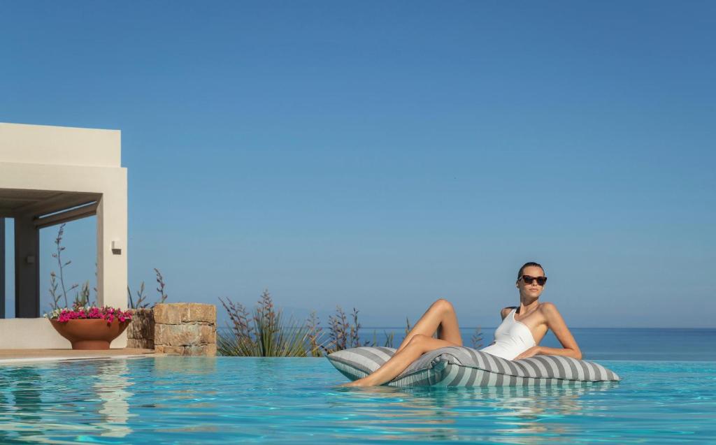 Sieviete atpūšas uz peldoša spilvena baseinā, baudot saulainu laiku un skatu uz jūru. Fons ir moderns arhitektūras elements ar ziedu podu.