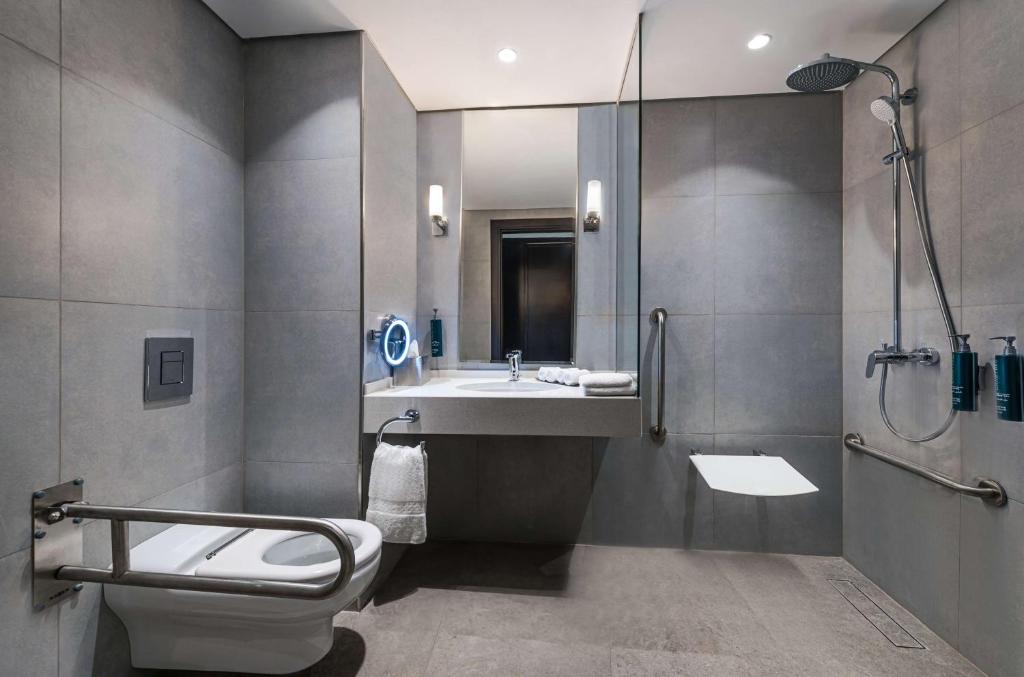 Mūsdienīgs vannas istabas interjers ar pelēkām flīzēm, dušas zonu un piekļuves gultni. Uz galda redzami dvieļi un higiēnas līdzekļi, kas rada ērtu atmosfēru.