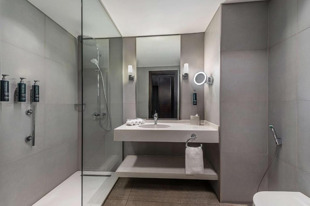 Mūsdienīgs vannas istabas interjers ar stikla dušu, pelēkiem flīzēm un elegantu izlietni. Vannas istabā ir pieejami higiēnas līdzekļi un dvieļi.