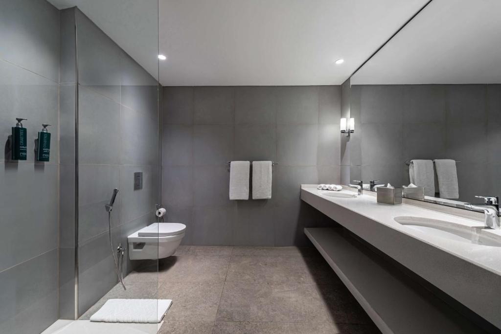Mūsdienīgs vannas istabas interjers ar pelēkām flīzēm, elegantu izlietni un higiēnas piederumiem uz plaukta. Gaismas elementi piešķir telpai siltumu un komfortu.