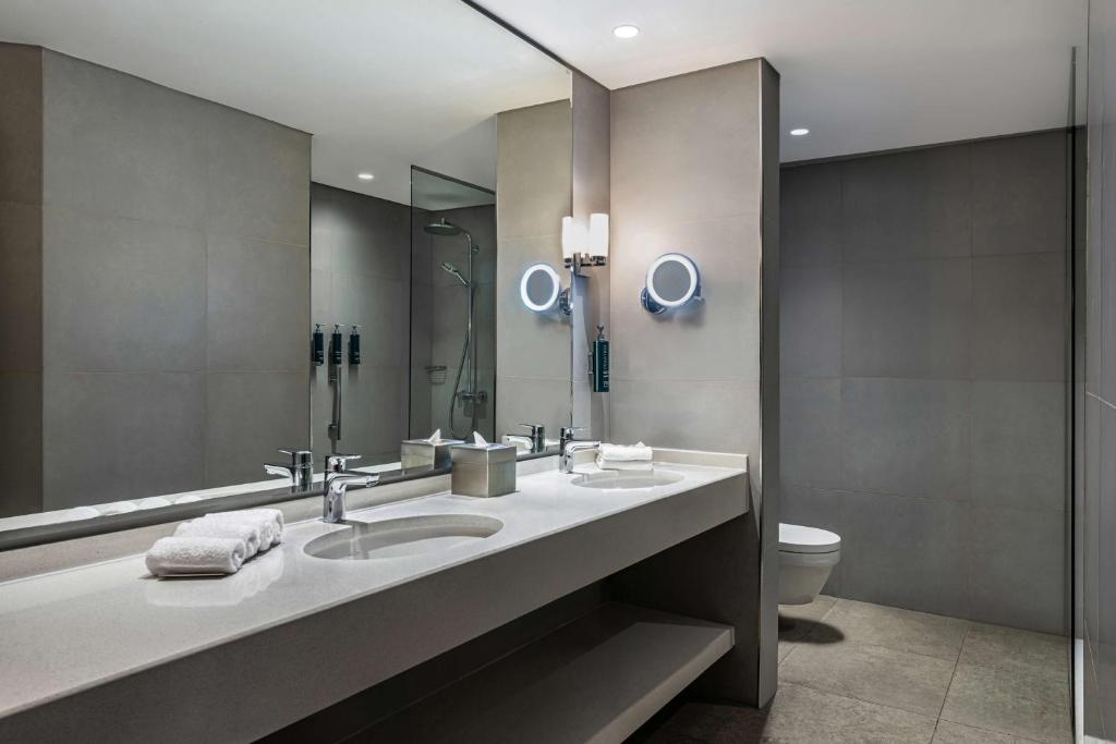 Moderns vannas istabas interjers ar divām izlietnēm un spoguli, kas rada elegantu un funkcionālu atmosfēru. Gaišas flīzes un mūsdienīgi apgaismojumi papildina kopējo dizainu.