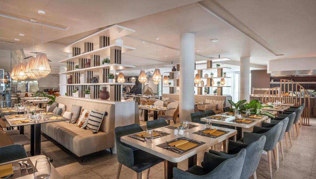 Mūsdienīgs restorāna interjers ar elegantiem galdiem un krēsliem, apgaismots ar stilīgām lampām. Telpa ir iekārtota ar zaļumiem un dekoratīviem elementiem, radot patīkamu atmosfēru.