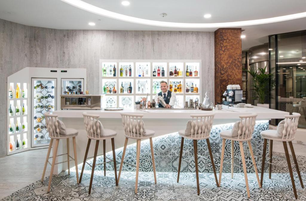 Mūsdienīgs bārs ar elegantu interjeru, baltiem krēsliem un plašu dzērienu izvēli. Apskatāmas glāzes un alkohola pudeles, kas izvietotas stilīgā vitrīnā.