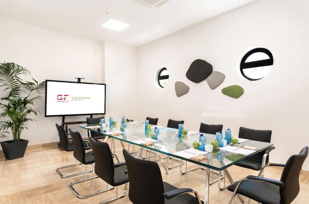 Konferenču telpa ar modernu dizainu, stikla galdu un melnām krēslām, kurā redzams ekranizēts zīmols "GF Hoteles Victoria". Telpā ir arī dekoratīvas augu elementu un sienu dekori.
