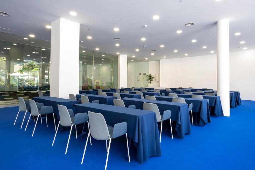 Konferenču telpa ar zilām galdautām un pelēkām krēsliem, plašs interjers ar gaišu apgaismojumu un stikla sienām. Ideāli piemērota sanāksmēm un pasākumiem.