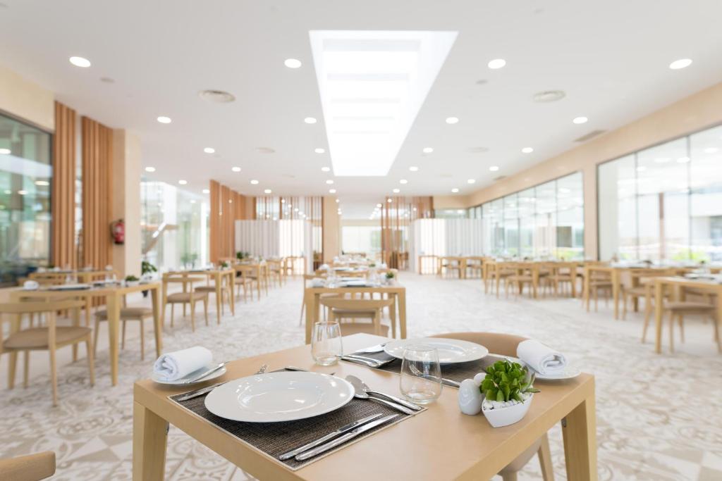 Mūsdienīgs restorāns ar gaišu interjeru, kurā redzami koka galdi un krēsli, kā arī eleganti galda piederumi. Lielas logu virsmas nodrošina dabīgu apgaismojumu.