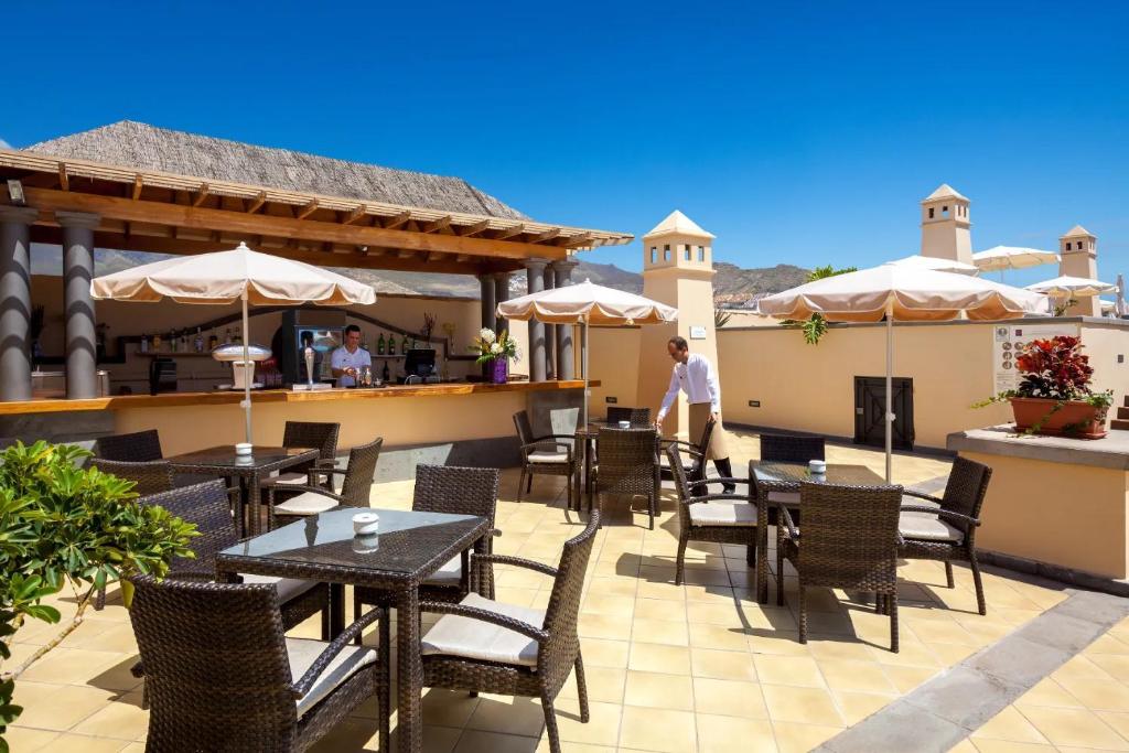 Skaists terases bārs ar saulessargiem un ērtām mēbelēm, kur apkalpo darbinieks. Fons ar skaidru zilu debesu un kalnu ainavu.