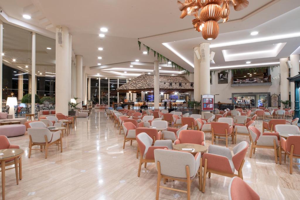 Mūsdienīga viesnīcas atpūtas telpa ar elegantiem, rozā un pelēkiem krēsliem un koka galdiem. Telpā ir daudz dabīgās gaismas un stilīgs apgaismojums, radot patīkamu atmosfēru.