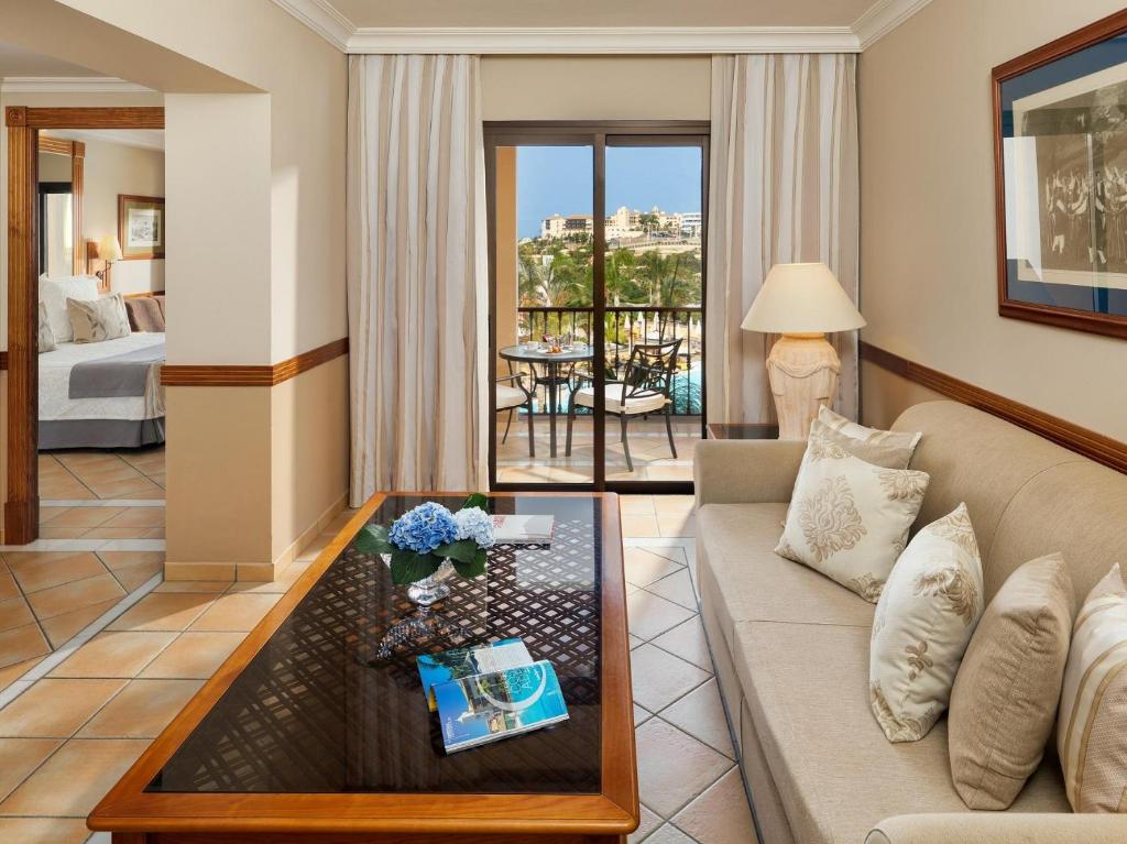 Mūsdienīgs viesnīcas numurs ar viesistabu, kurā ir dīvāns un galdiņš ar ziediem, kā arī balkons ar skatu uz baseinu. Interjers ir gaišs un elegants, radot relaksējošu atmosfēru.