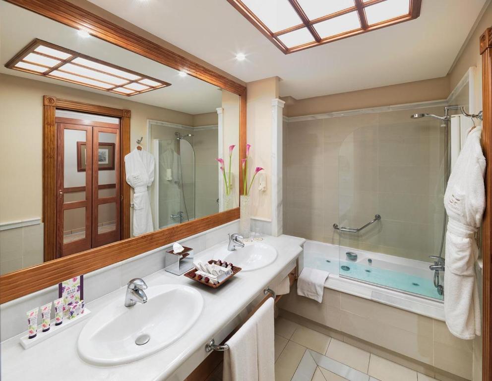 Elegants viesnīcas vannas istabas interjers ar divām izlietnēm, vannu un dušu. Svaigas ziedu kompozīcijas un gaismas elementi rada relaksējošu atmosfēru.