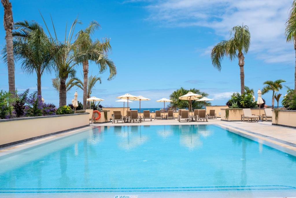 Peldbaseins ar palmām un sauļošanās krēsliem, skats uz jūru un zilo debesu fonu. Ideāla atpūtas vieta.