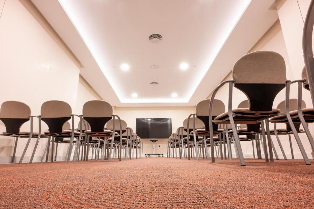 Konferenču telpa ar rindu krēsliem, kas vērsti pret ekrānu. Telpa ir gaiša, ar modernu apgaismojumu un siltu paklāju.