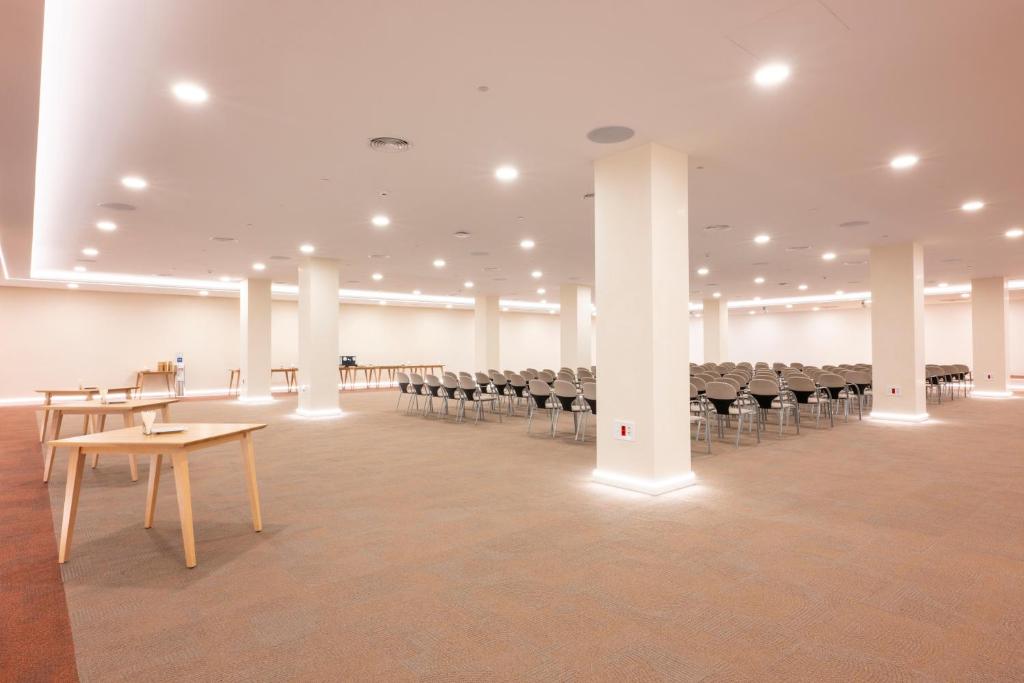 Plaša konferences zāle ar gaišu interjeru, kurā redzami galdi un rindas krēslu. Apgaismojums ir mīksts un moderns, radot patīkamu atmosfēru pasākumiem.