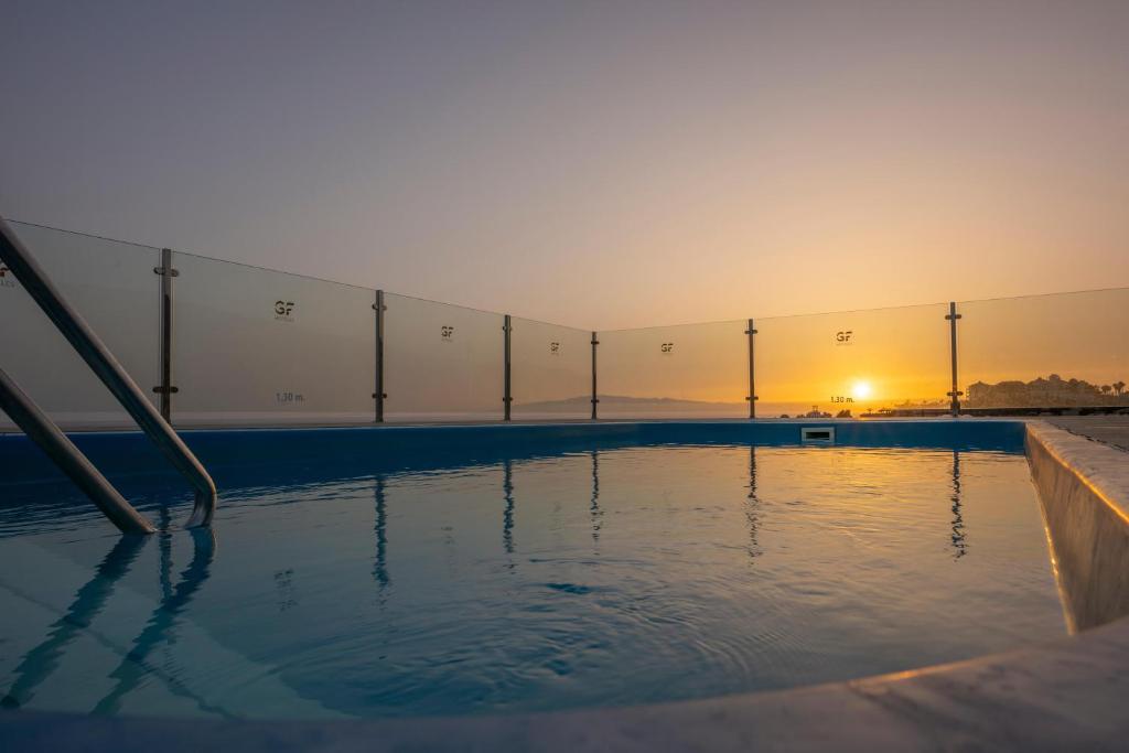 Sauļošanās baseins ar stikla barjeru, kas piedāvā skatu uz saulrietu. Rāmā ūdens virsma atspoguļo apkārtējo ainavu.