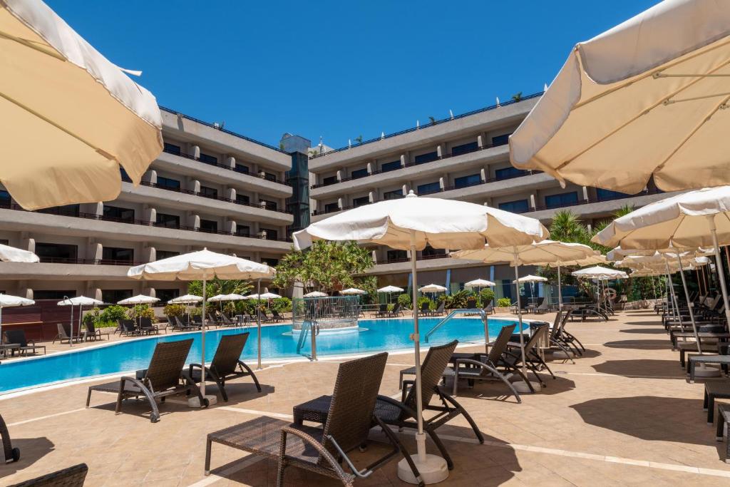 Viesnīcas baseins ar sauļošanās krēsliem un saulessargiem, apkārtne ir apzaļumota. Skats uz modernu viesnīcas ēku, skaidra debess.