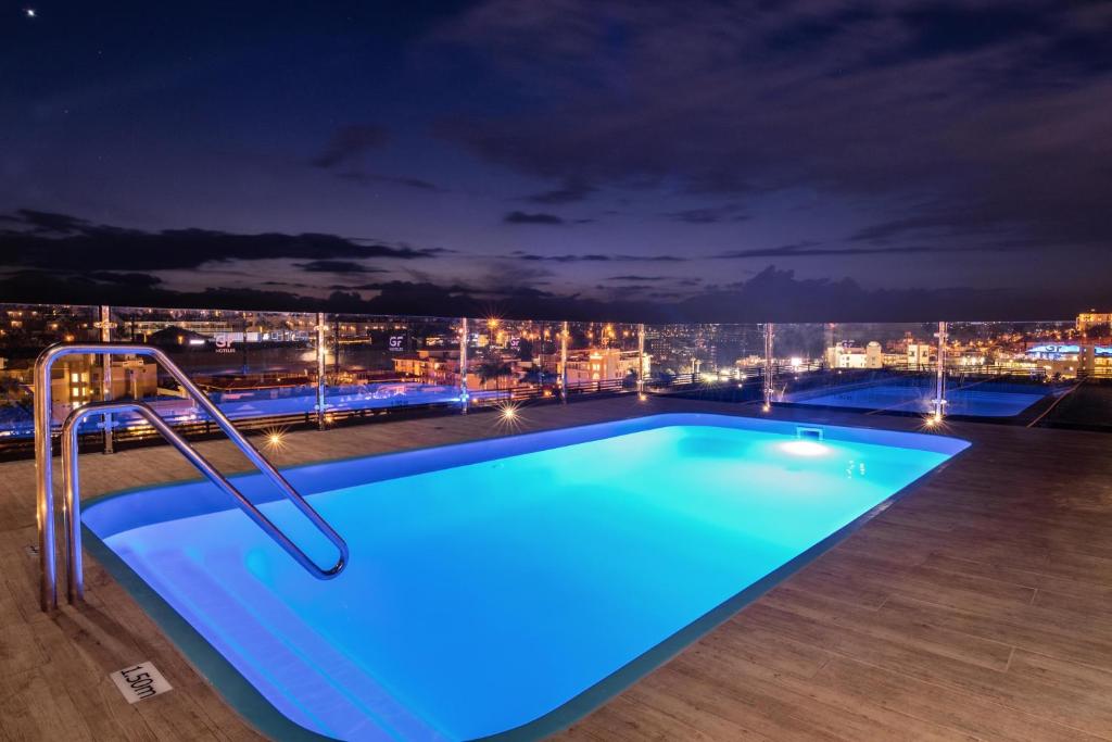 Mūsdienīgs peldbaseins ar zilganu apgaismojumu, novietots uz jumta, ar skatu uz pilsētas apgaismojumu naktī. Tuvumā redzama stikla barjera un moderns koka grīdas segums.