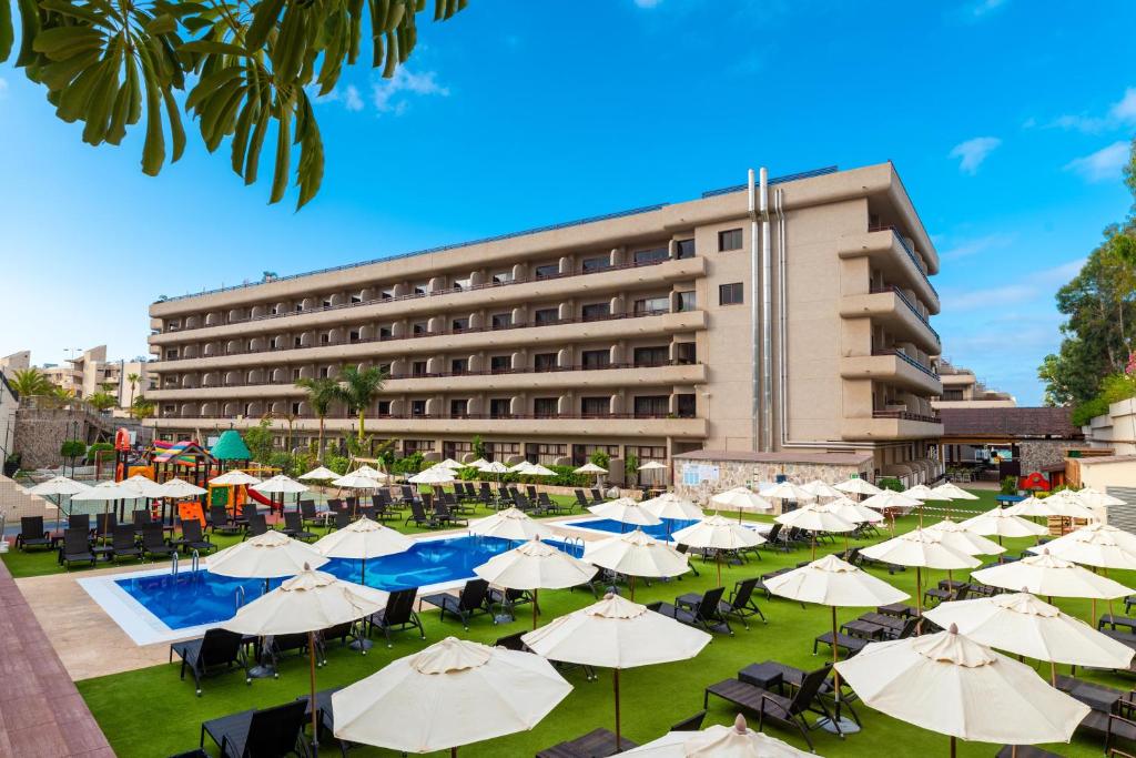 Viesnīcas ārpuse ar baseinu un sauļošanās krēsliem, apkārt palmu un saulessargu ainava. Ideāla vieta atpūtai un izklaidei ģimenēm.