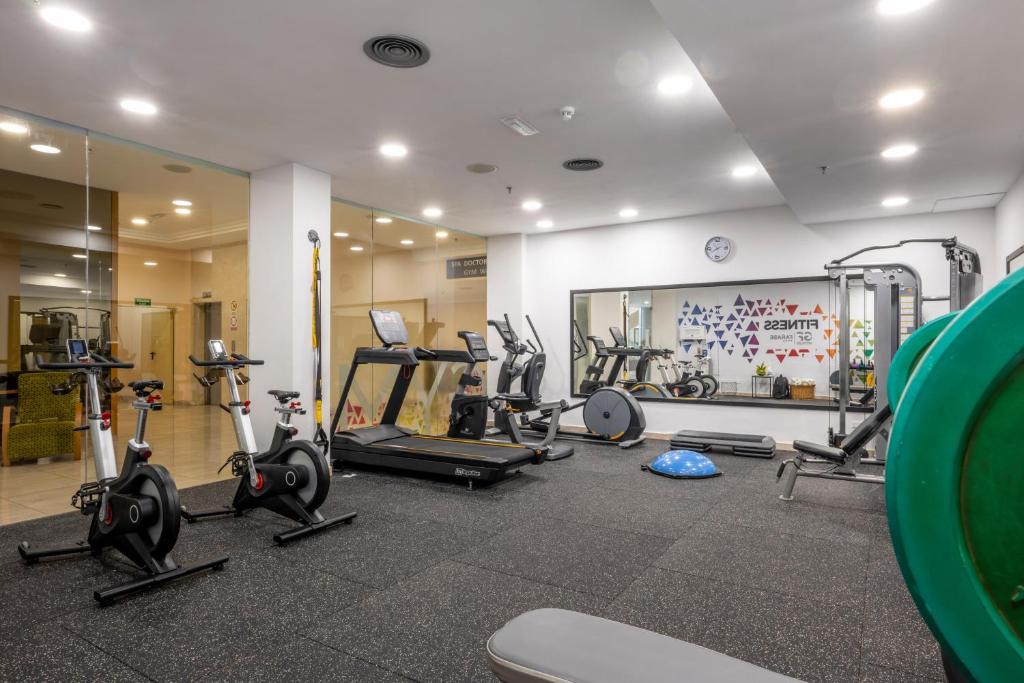 Mūsdienīga sporta zāle ar dažādām trenažieru ierīcēm, tostarp velotrenažieriem, skrejceliņu un svaru iekārtām, kas izvietotas uz melnas gumijas grīdas. Sienā redzama dekoratīva krāsaina apdare.