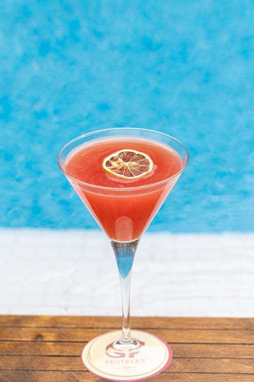 Sarkans kokteilis ar žāvētu citrona šķēli, novietots uz koka virsmas ar zilu baseina fonu.