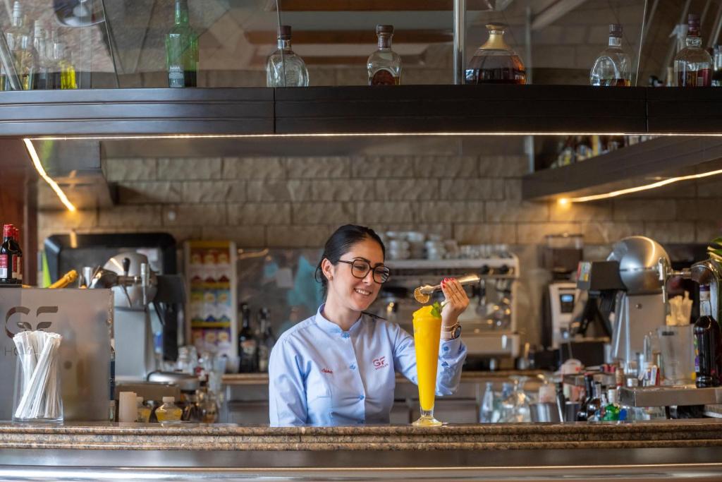 Bāra darbiniece, kas pasniedz dzērienu, smaidot, kamēr aiz viņas redzama bāra aprīkojums un dzērieni.