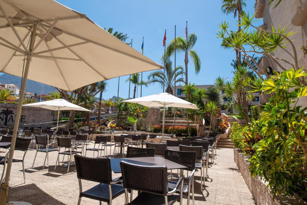 Āra kafejnīca ar saulessargiem un galds, apzaļumots ar palmām un ziediem, ideāla vieta relaksācijai. Skats uz viesnīcu un kalniem, saulaina atmosfēra.