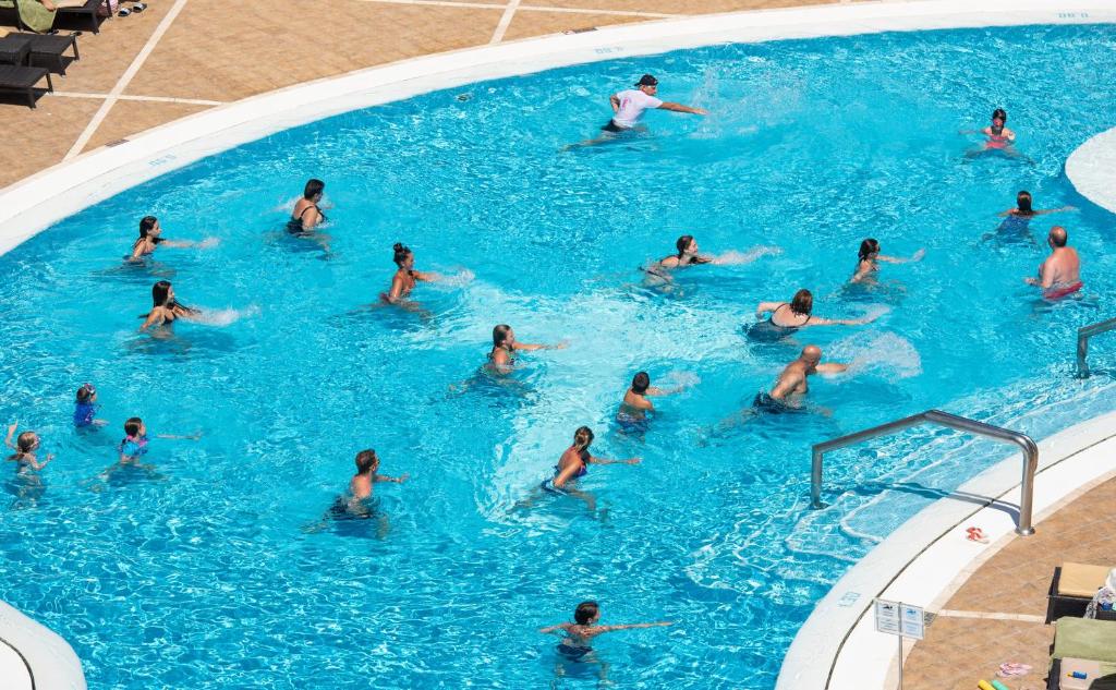 Baseinā notiek grupas peldēšanas nodarbība, kurā piedalās gan pieaugušie, gan bērni, izklaidējoties ūdenī. Visi apmeklētāji izskatās priecīgi un aktīvi.