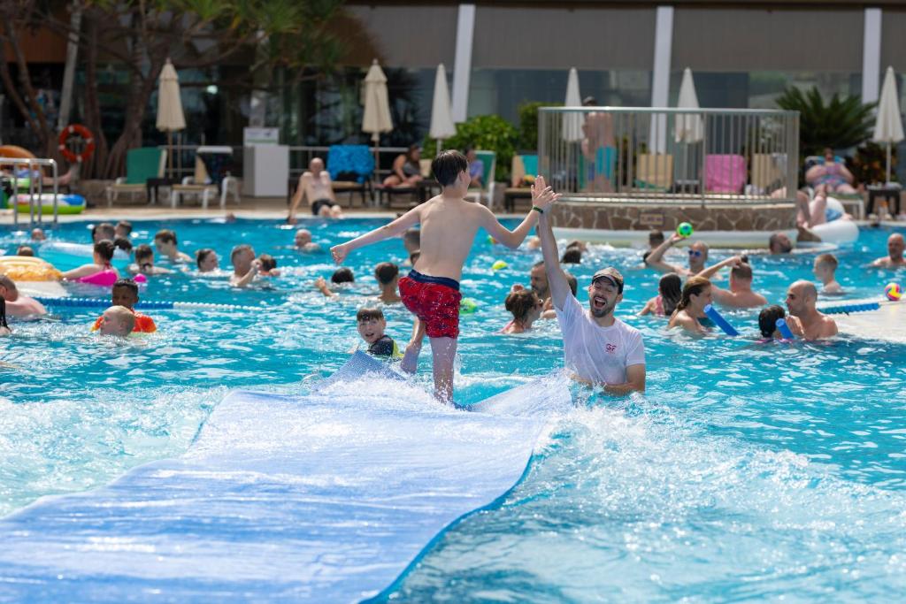 Bērni izklaidējas pie baseina, pārvietojoties pa slideno virsmu, kamēr citi peld un atpūšas ūdenī. Saule spīd un atmosfēra ir dzīvīga un priecīga.
