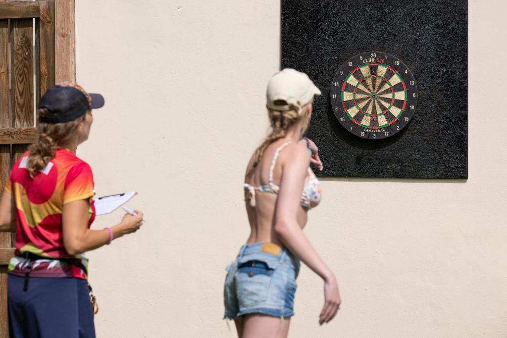 Divas personas spēlē dartus, viena no tām gatavojas mest bultu mērķī, kas piestiprināts pie sienas. Otrā persona pieraksta rezultātus.