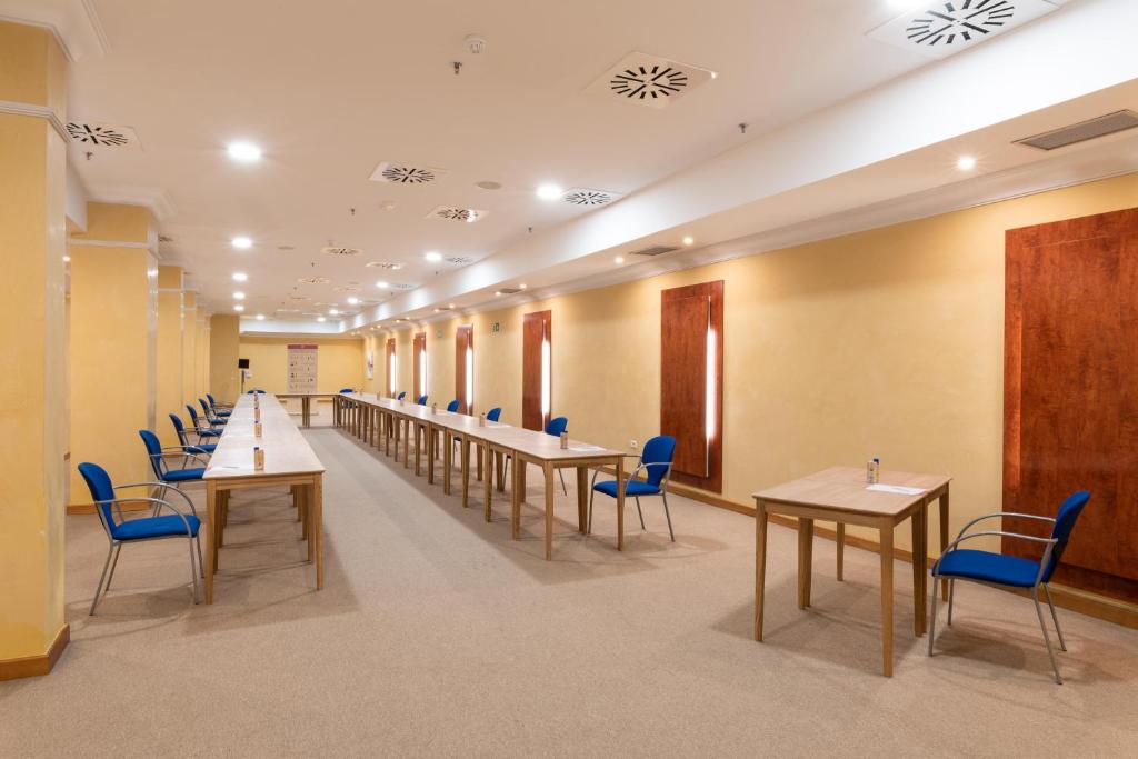 Konferences zāle ar garām galdiem un zilu krēslu rindām, spilgti apgaismota un ar dzeltenām sienām.