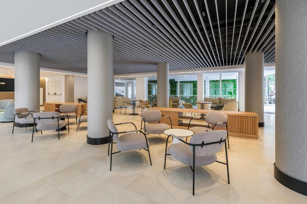 Mūsdienīgs viesnīcas vestibils ar elegantiem krēsliem un koka mēbelēm, kas rada patīkamu un relaksējošu atmosfēru. Gaisma un zaļumi papildina interjera dizainu.