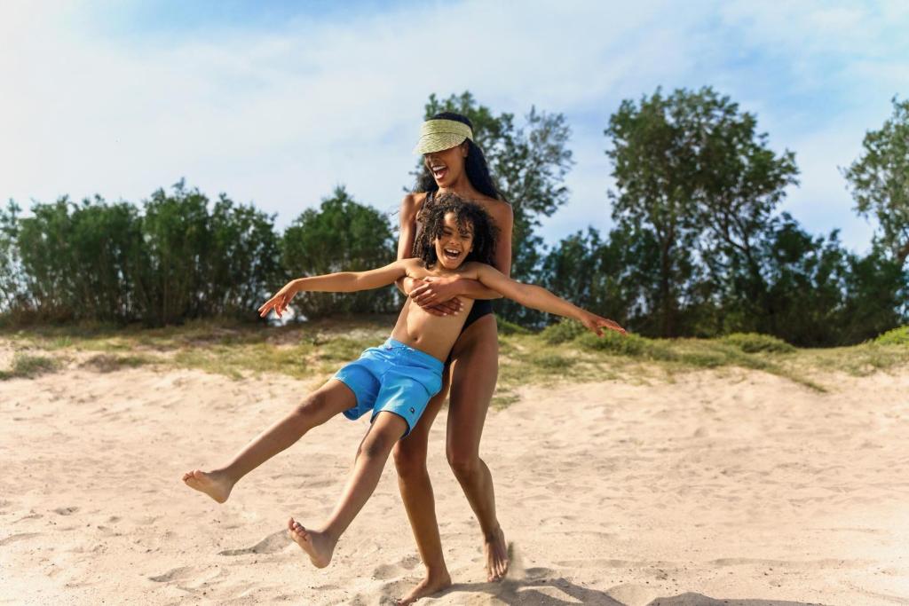 Māte un dēls priecīgi izbauda laiku pludmalē, kur viņa nes viņu, kamēr abi smejas. Apkārt ir smilšaina vide un zaļi krūmi.
