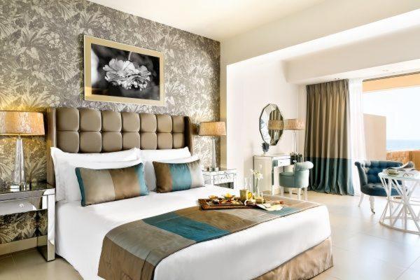 Mūsdienīgs un elegants viesnīcas numurs ar plašu gultu, greznu dekoru un skatu uz jūru, kas piedāvā relaksējošu atmosfēru. Uz galda redzams brokastu plate ar gardumiem un ziediem.