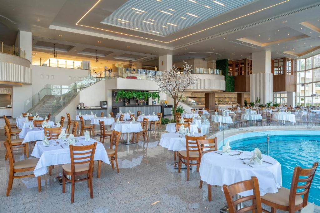 Liels un elegants restorāns ar baltām galdautām un koka krēsliem, kurā ir redzams iekšējais baseins un zaļumi.