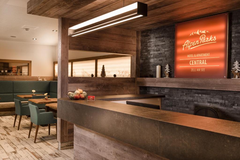 Mūsdienīgs viesnīcas reģistratūras interjers ar koka apdari un ērtām mēbelēm, kurā redzams "AlpenParks" viesnīcas zīme. Siltas krāsas un patīkama atmosfēra veido viesnīcas centrālo daļu.