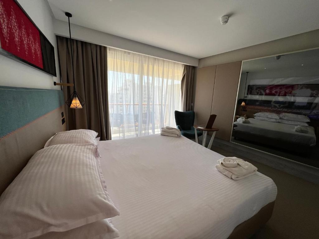 Mūsdienīga viesnīcas numura iekšskats ar plašu gultu, stilīgu apgaismojumu un ērtu atpūtas krēslu. Liels logs ļauj ieplūst dabīgai gaismai, radot mājīgu atmosfēru.