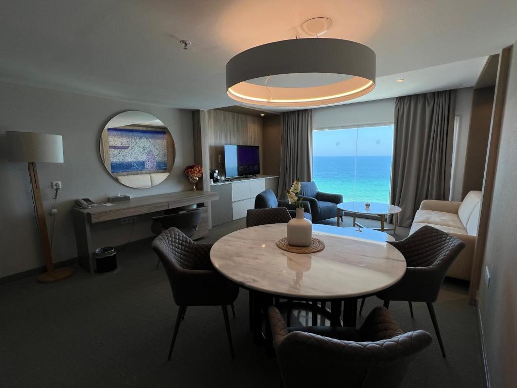 Mūsdienīga viesnīcas numura interjers ar panorāmas skatu uz jūru, iekārtots ar ērtu dīvānu, galdu un televizoru. Interjerā ir eleganti akcenti un gaismas elementi.