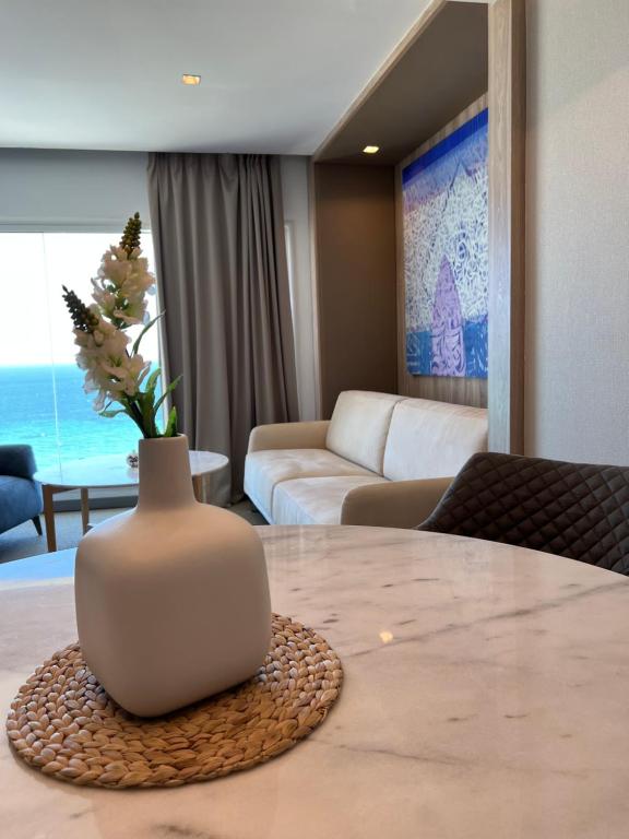 Mūsdienīgs viesistabas interjers ar jūras skatu, baltu dīvānu un dekoratīvu vāzi uz galda. Gaismas tonis un mākslas darbs uz sienas piešķir telpai eleganci.