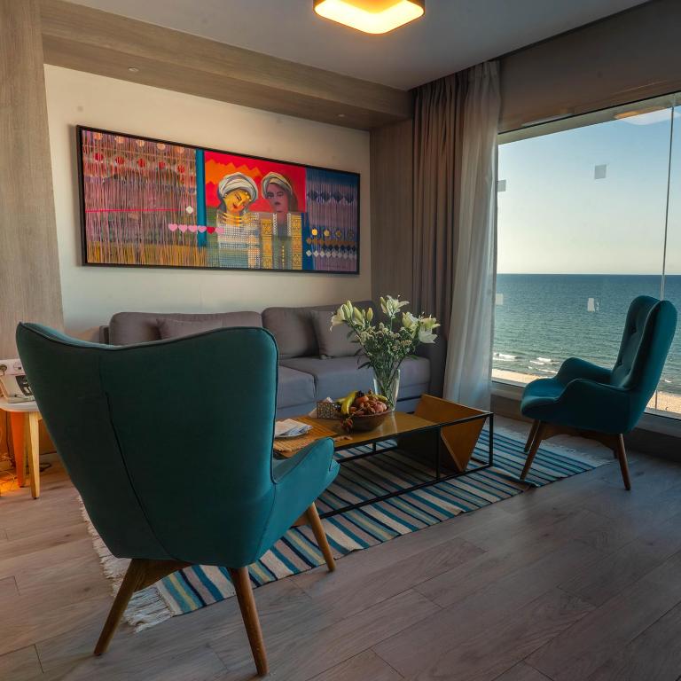 Mūsdienīgs viesistabas interjers ar jūras skatu, ko rotā krāsaina glezna un stilīgas mēbeles. Galdā novietots ziedu pušķis un augļi, radot mājīgu atmosfēru.