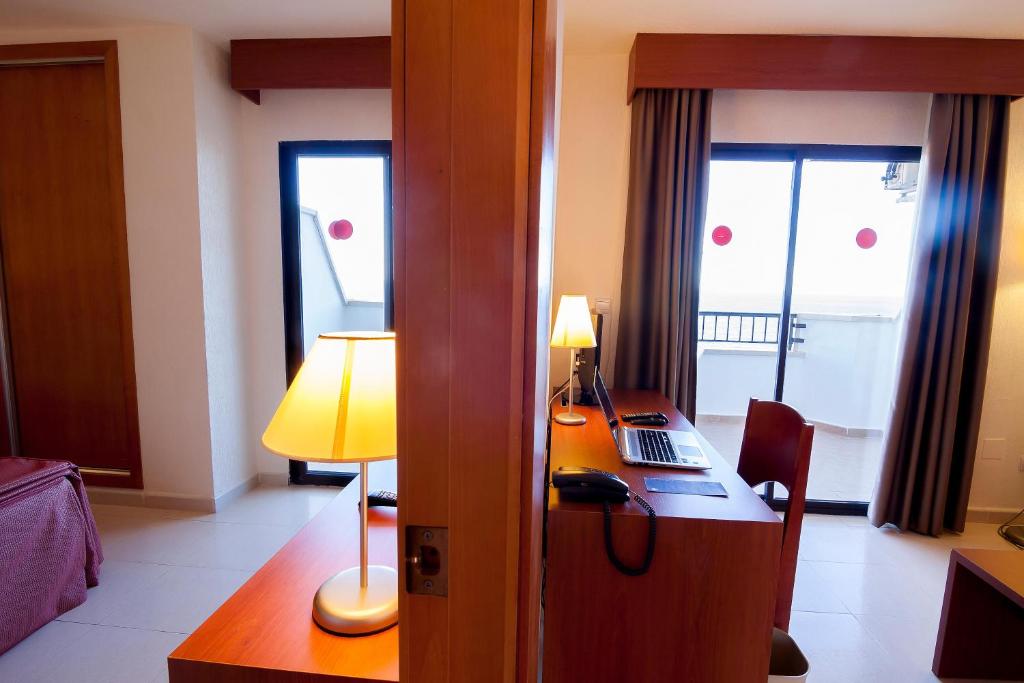 Mūsdienīgs viesnīcas numurs ar darba galdu, lampu un logu, caur kuru redzams balkons. Interjers ir gaišs un ērts, radot patīkamu atmosfēru.