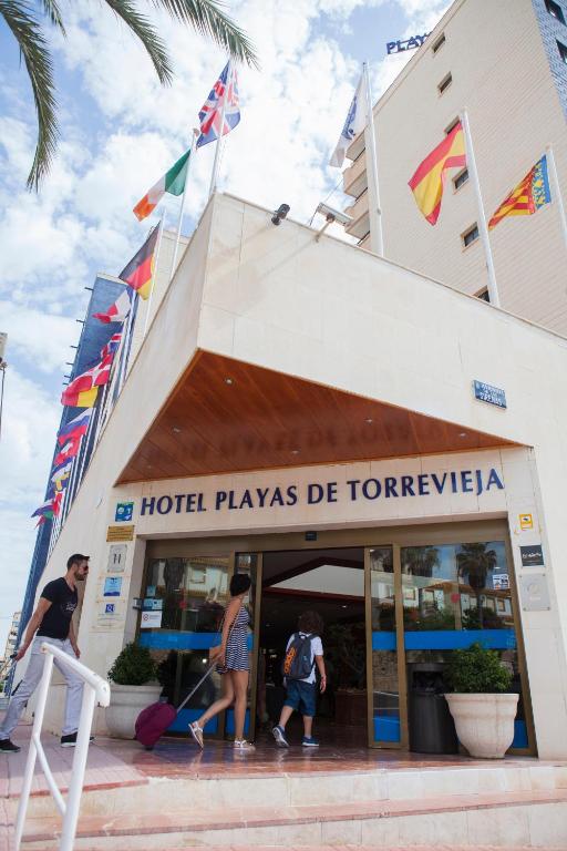 Ieejas vārti uz viesnīcu "Playas de Torrevieja", kas atrodas pie pludmales, ar karogiem no dažādām valstīm. Apmeklētāji ienāk viesnīcā, kas piedāvā komfortu un relaksāciju.