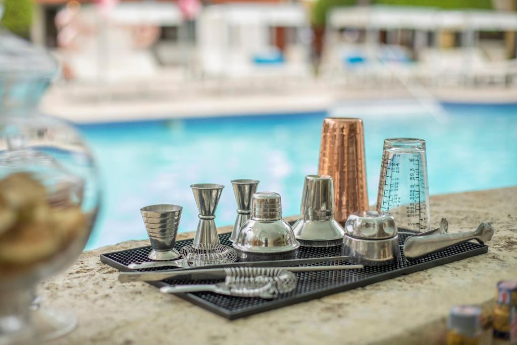 Metāla kokteiļu instrumenti, tostarp mērkausi un kratītāji, novietoti uz melna palikņa blakus baseinam.