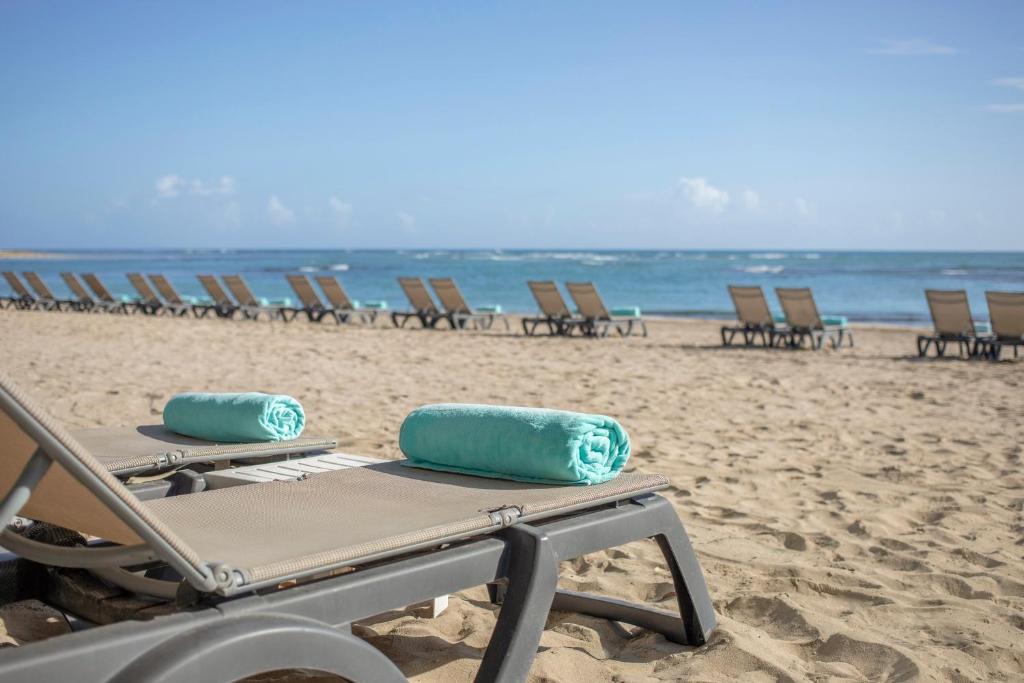 Pludmales atpūtas krēsli ar ziliem dvieļiem, kas izvietoti smilšu pludmalē, ar skatu uz mierīgu jūru un debesīm.