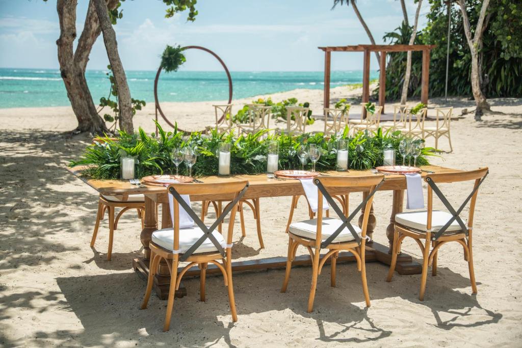 Pludmales svinību galds ar zaļumiem un traukiem, kas novietots pie jūras. Apkārt ir koka krēsli un dekorācijas, radot elegantu atmosfēru.
