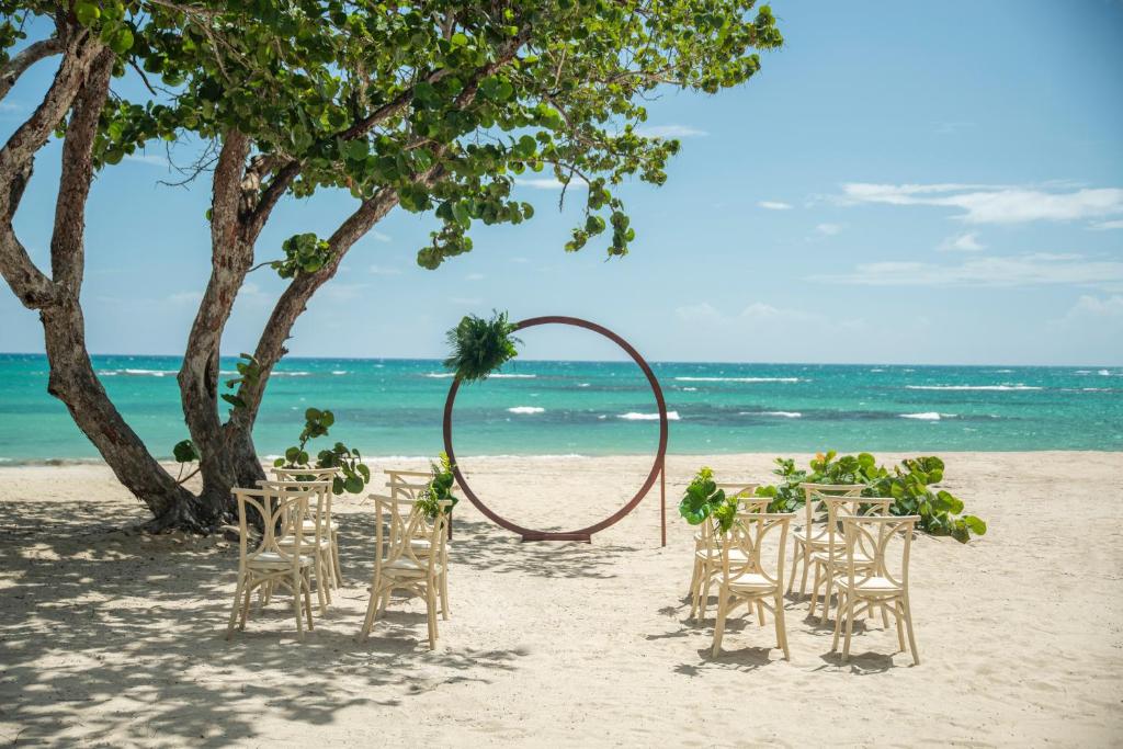 Pludmales kāzas ar koka krēsliem un dekoratīvu loku, kas rotāts ar zaļumiem, fons ir jūra un debesu zilums.