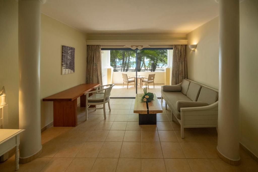 Moderns viesnīcas numurs ar gaišām sienām un mēbelēm, kurā ir dīvāns, galds un logi ar skatu uz dārzu. Interjers ir ērts un mājīgs, radot relaksējošu atmosfēru.