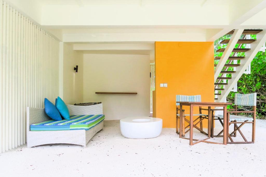 Mūsdienīgs dzīvojamais telpa ar baltu sienu, gaiši oranžu akcentu un dabīgām koka mēbelēm, kas ietver dīvānu un ēdamgaldu. Telpā ir ērti spilveni un gaiša atmosfēra.